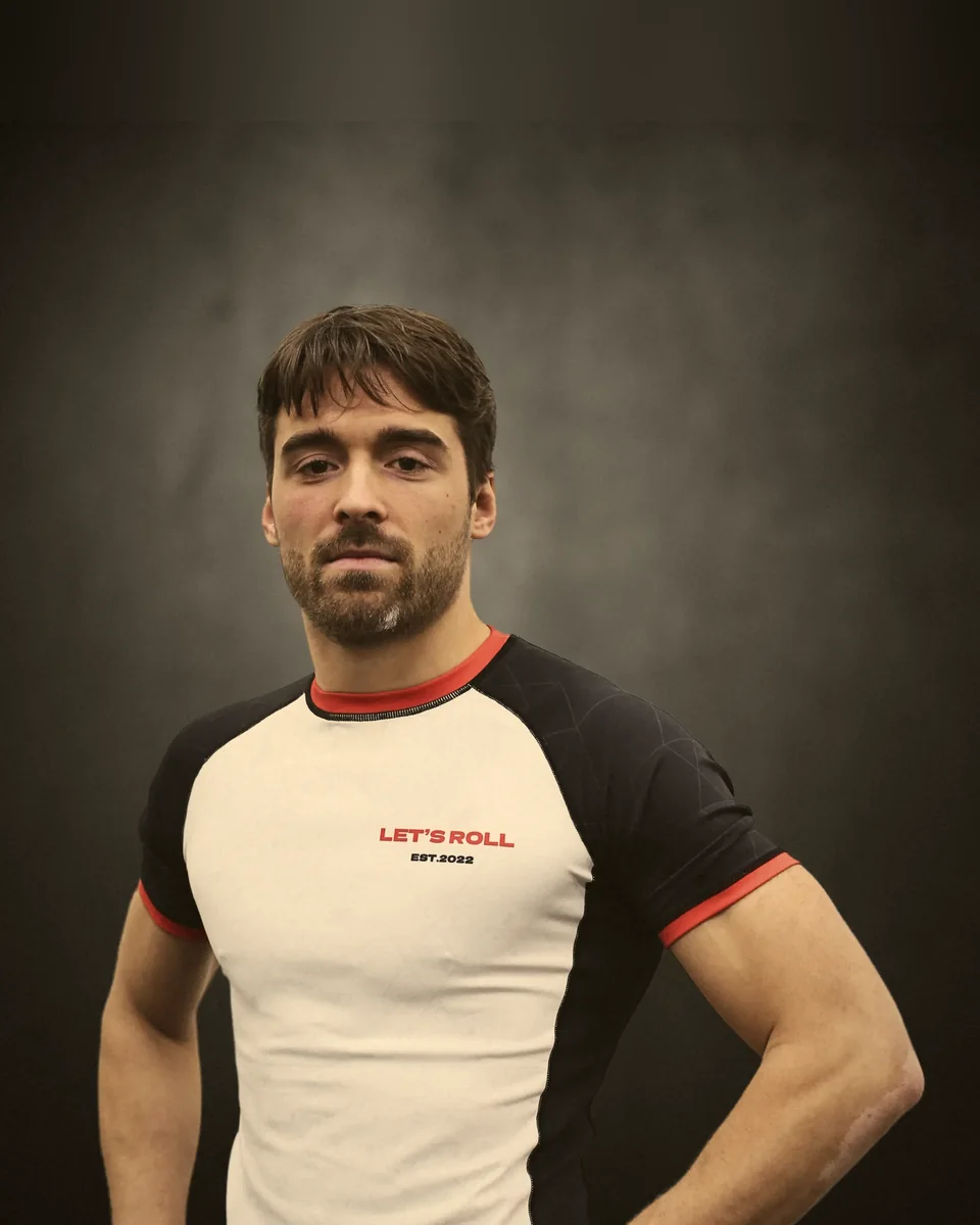 Coach der Let's Roll Academy im weiß-schwarzen Rashguard vor grauem Studiohintergrund.